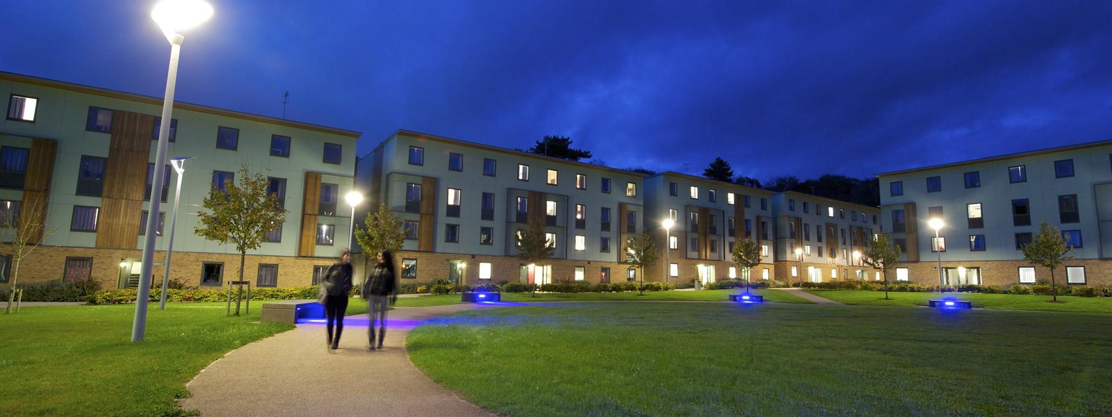 Two students walk along a path on campus at night with student accommodation in the background.