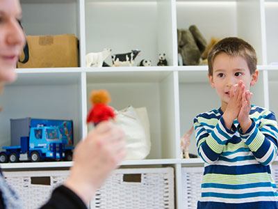 a small boy in front of shelves claps his hands at a lady holding a toy in the foreground