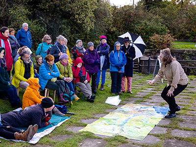 a group of people listen to someone talking and pointing at a large map on the ground