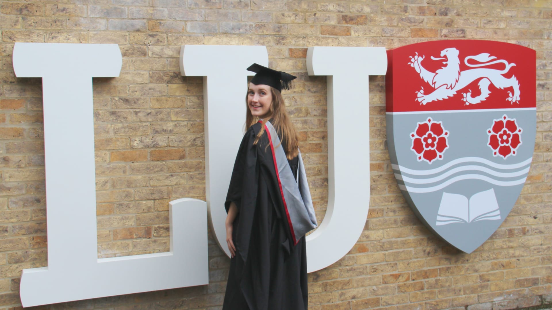 Catherine stands, wearing graduation robes, in front of a large sign saying LU.