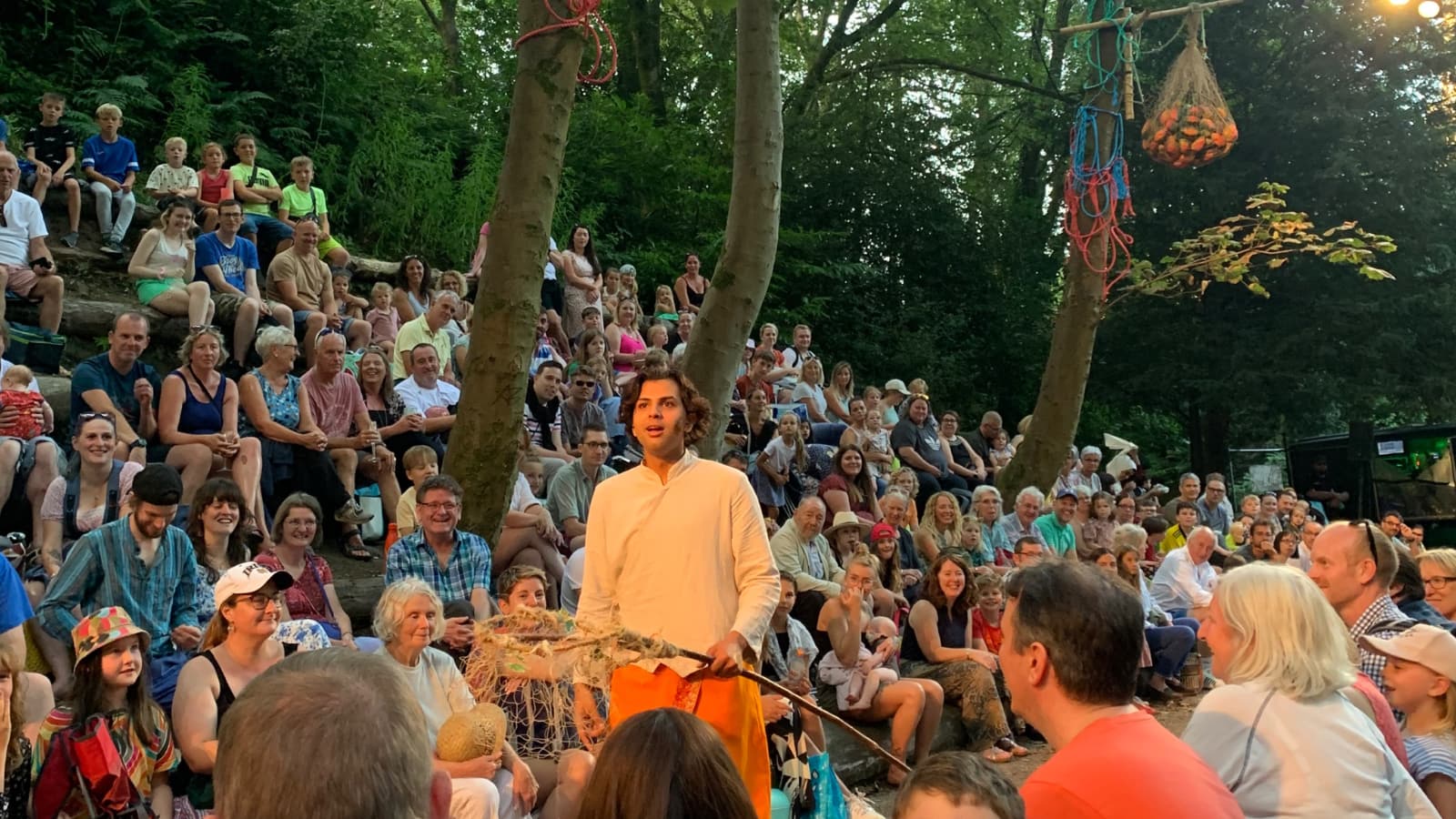 An actor playing Mowgli stands in a wooded glade surrounded by the audience