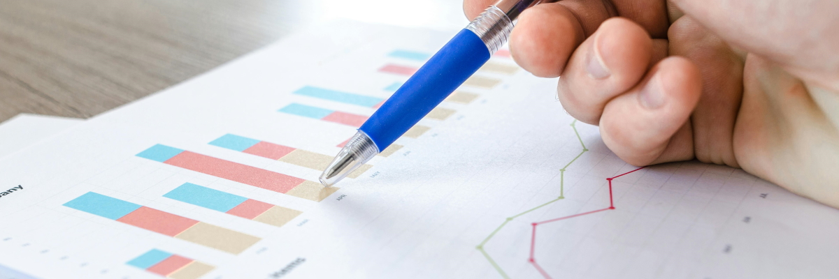 A hand holding a blue ballpoint pen pointing at bar charts and line graphs printed on white paper.