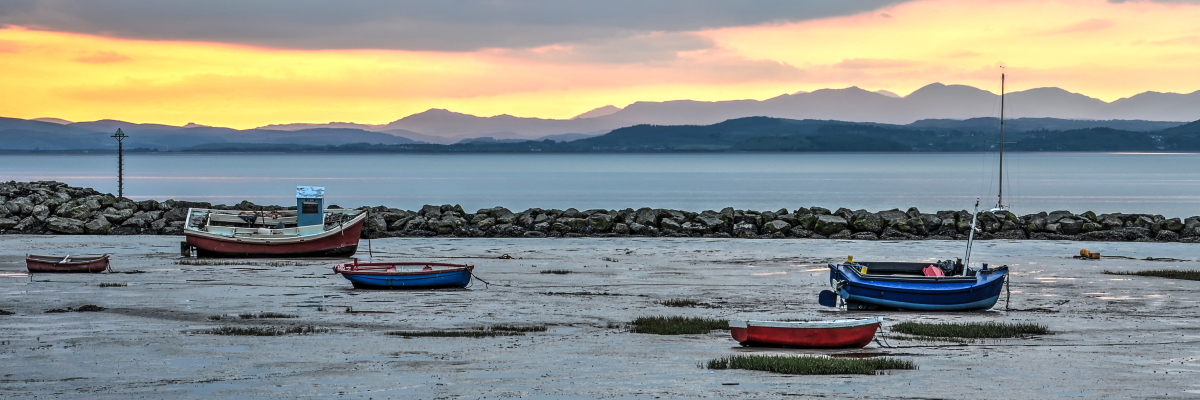 A group of small colourful fishing boats resting on the sand in Morecambe Bay at low tide at sunset.